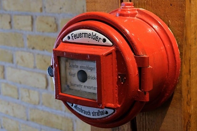 Why Coventry Businesses Need Advanced Intruder Alarm: A red fire alarm call point with German text is mounted on wood, highlighting the need for safety systems beyond just fire detection for businesses In Coventry.