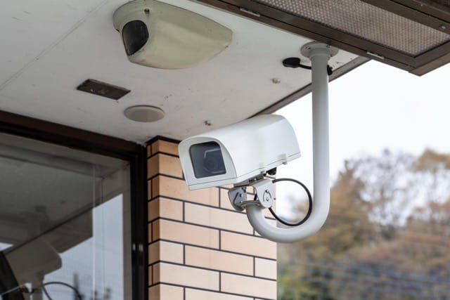 An outdoor bullet-style camera, part of Home Security Systems, is mounted on the corner of a brick building. Another dome camera is visible under the eaves above it.